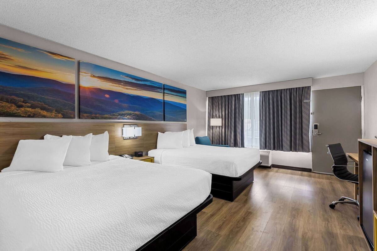 A hotel room with two beds, a desk, a chair, and a large painting of a sunset.