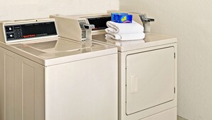 A front-loading commercial washer with a control panel, a folded white towel, and blue detergent boxes on top.