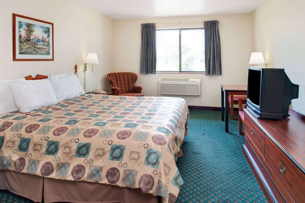 Ein Hotelzimmer mit einem großen Bett, einem Sessel, einem Fernseher und einem Fenster mit Vorhängen.