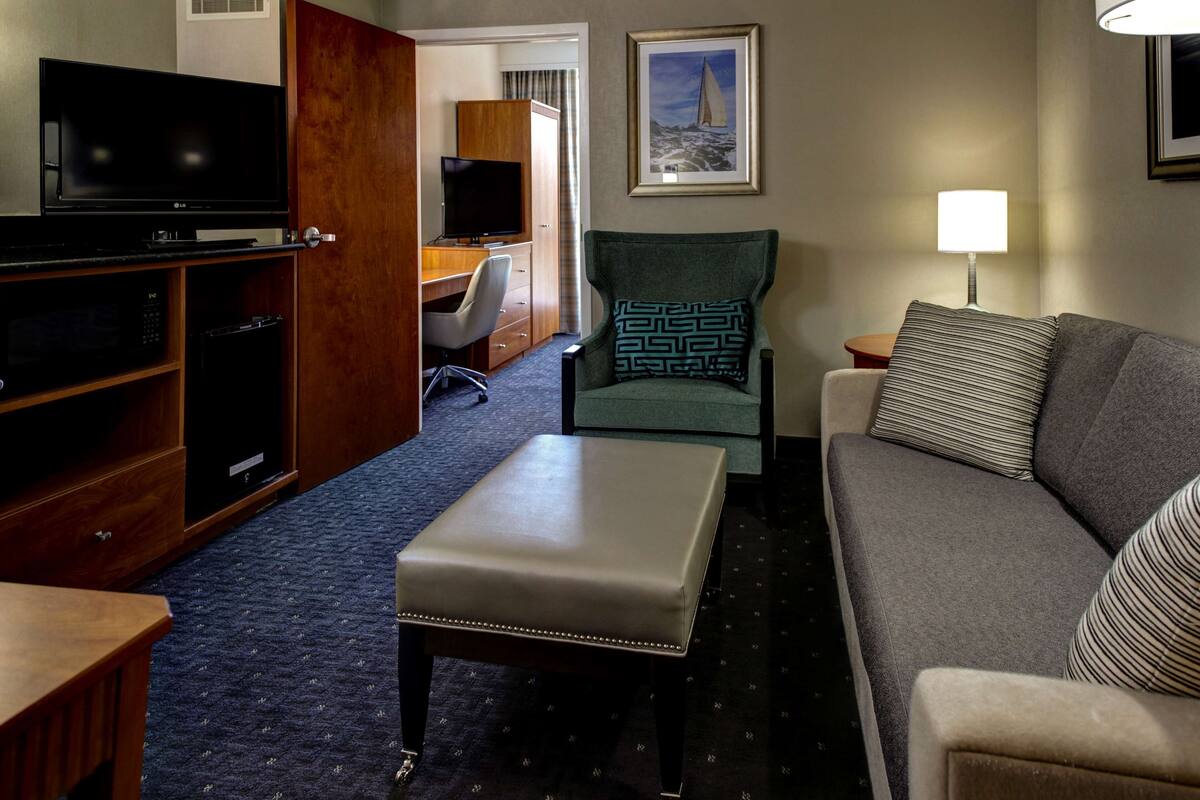 A hotel room with a TV, a desk, a green armchair, a grey sofa, and a lamp.