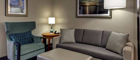 Habitación de hotel con un sofá gris, un sillón verde azulado, una mesita de noche con una lámpara y dos cuadros en la pared.