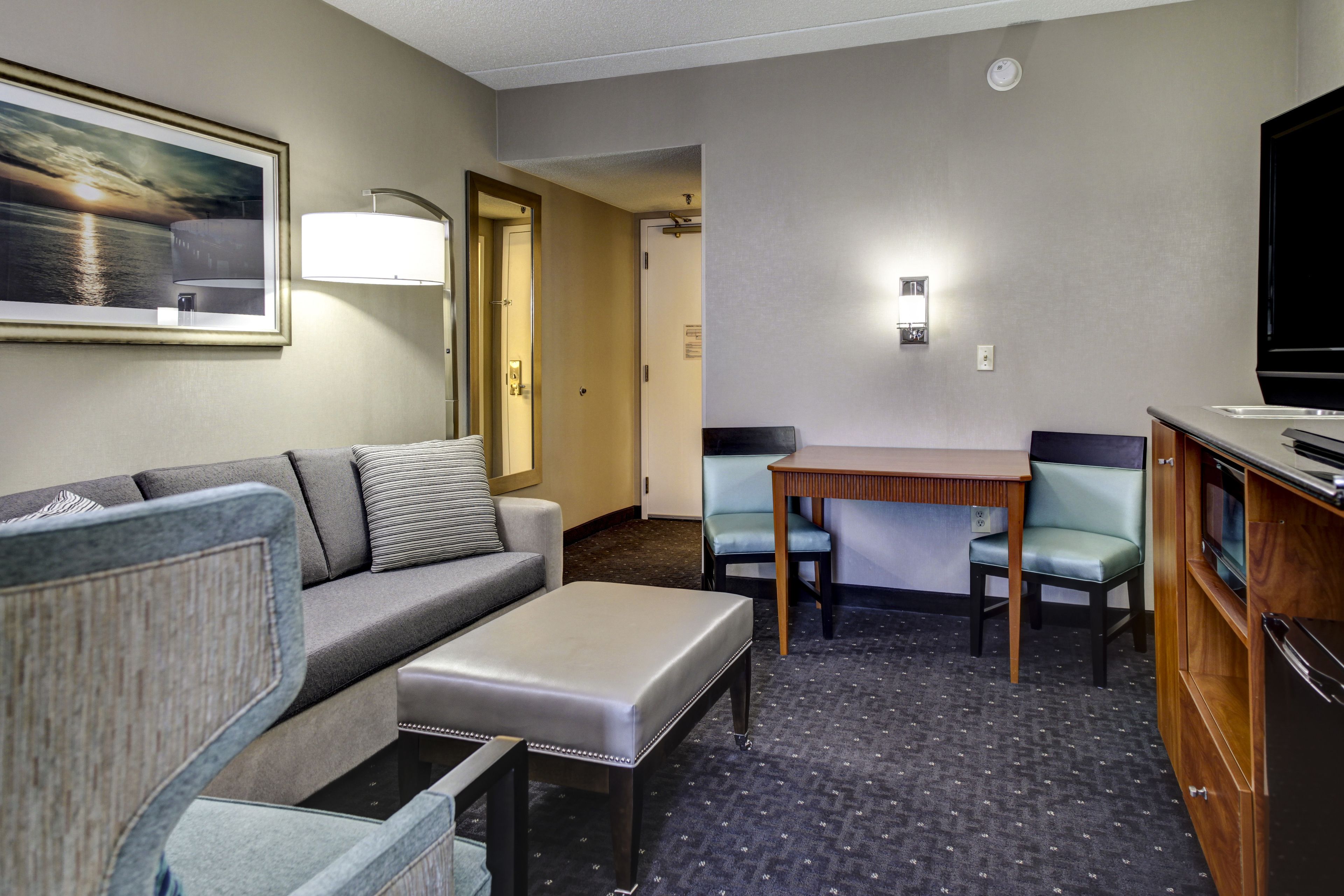 A hotel room with a sofa, ottoman, two chairs, a small table, a television, and a framed picture on the wall.