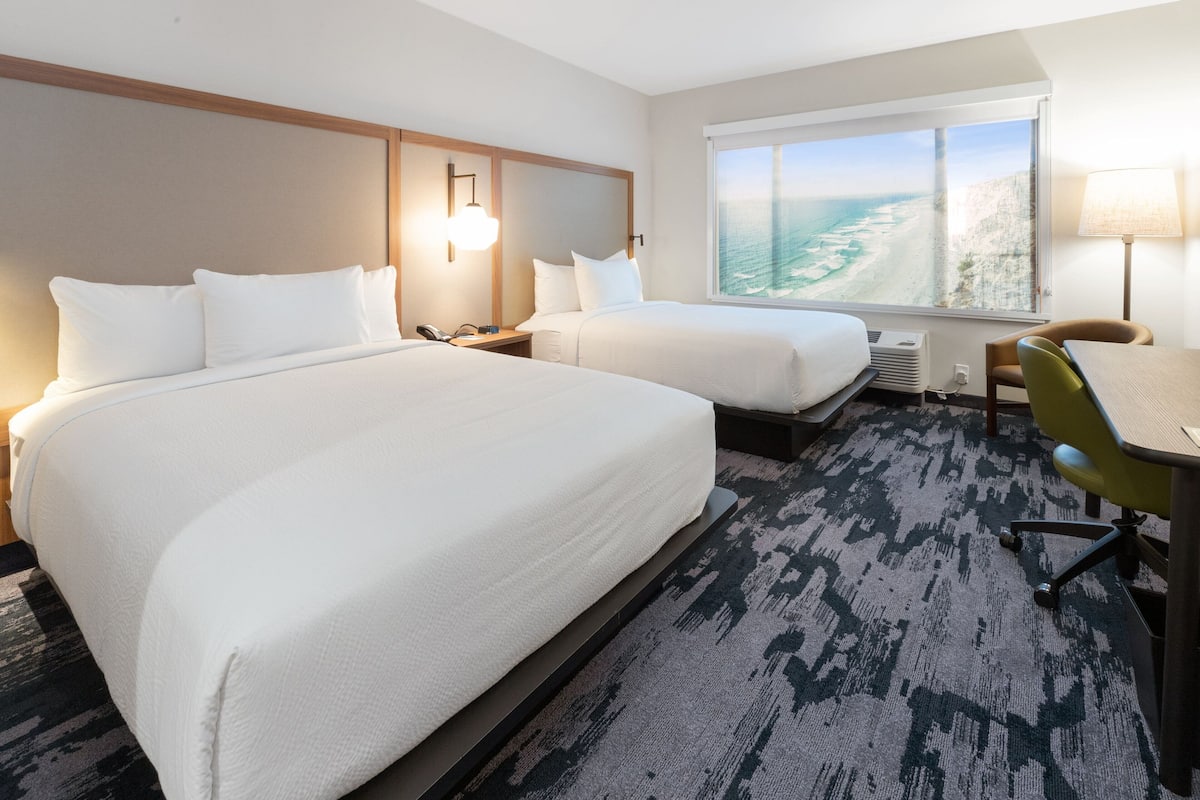 A hotel room with two beds, a desk, a chair, a window with a view of the ocean, and a lamp.