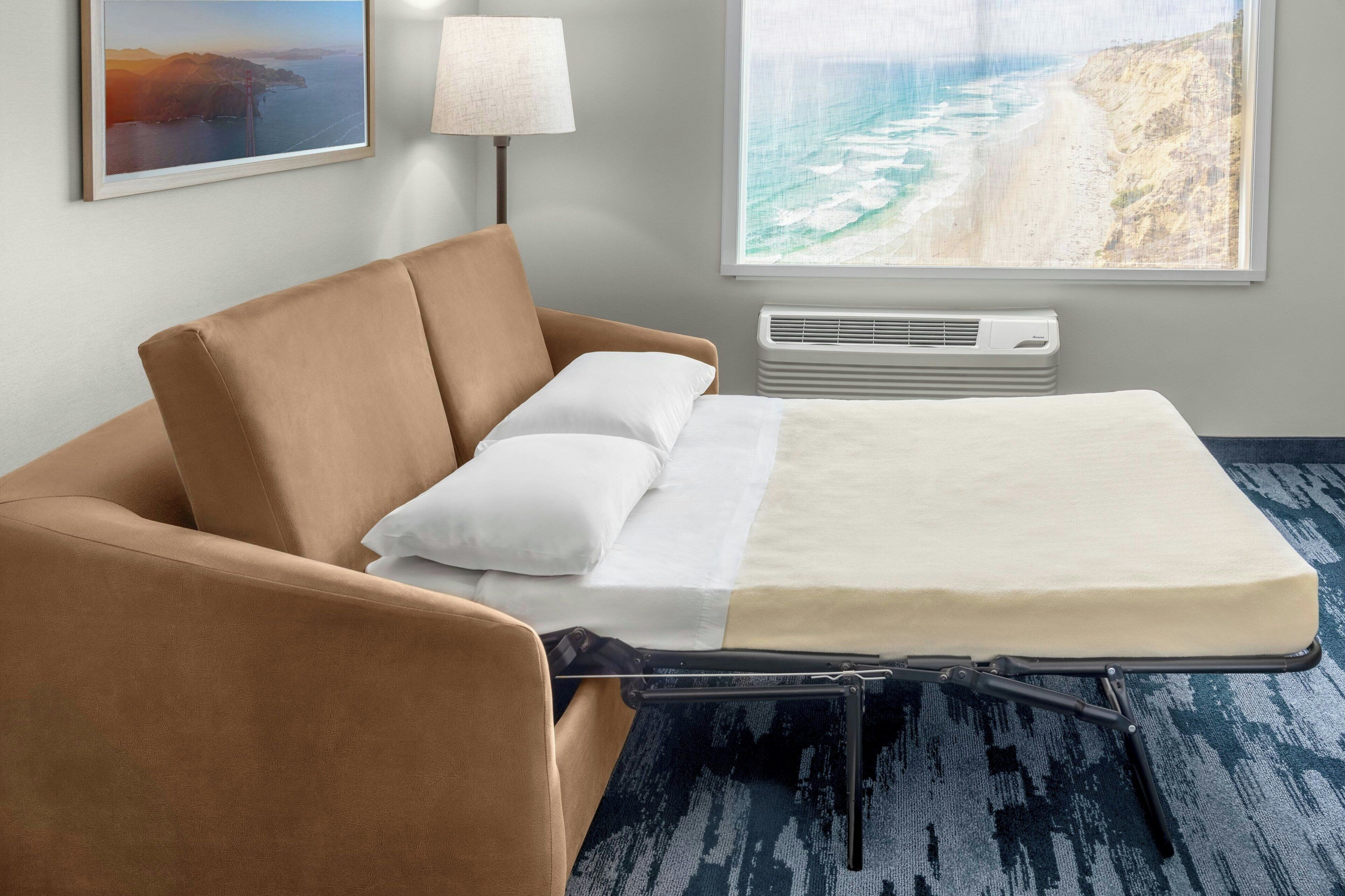 Habitación de hotel con cama, un sillón marrón, almohada blanca, lámpara y un ventanal con vista al mar.