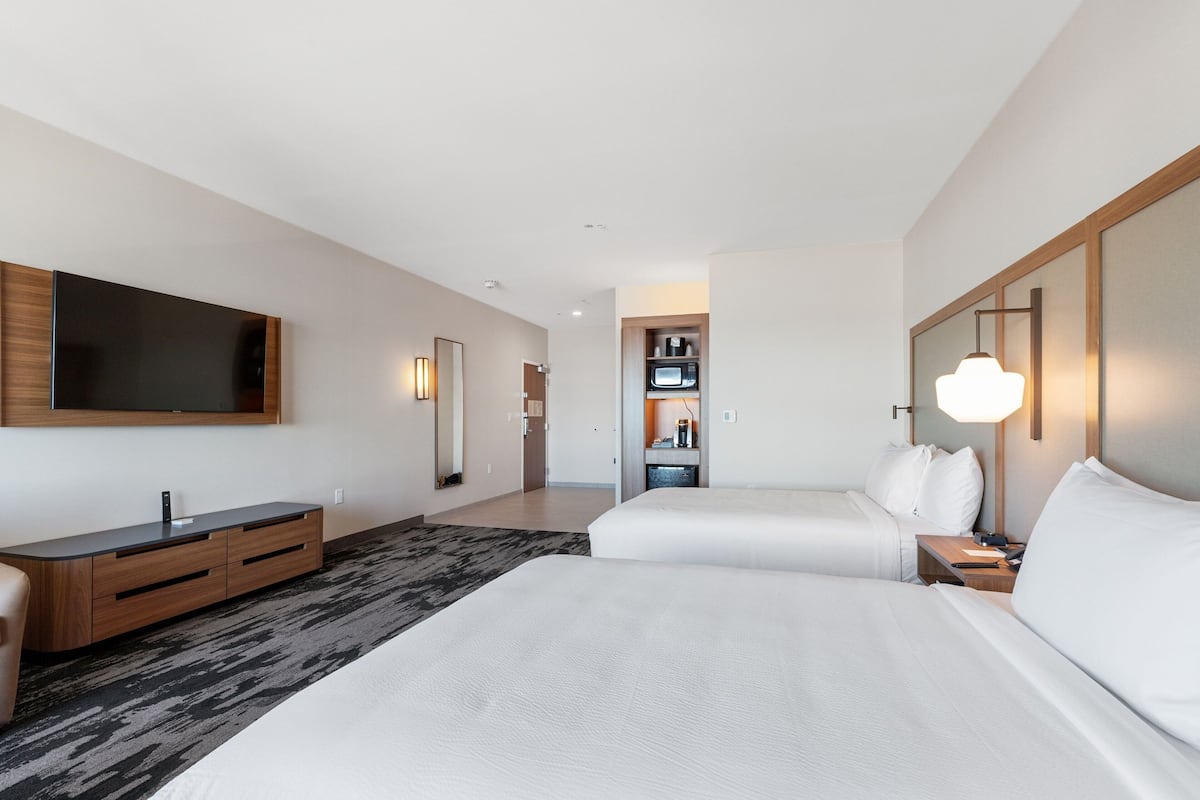 A modern hotel room with two beds, a flat-screen TV, a wooden console, a built-in microwave, and a bedside table with a lamp.