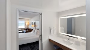 A hotel room with a bed, a bathroom with a large mirror, a sink, and a window. - Fairfield Inn & Suites by Marriott San Francisco Pacifica (Pacifica)
