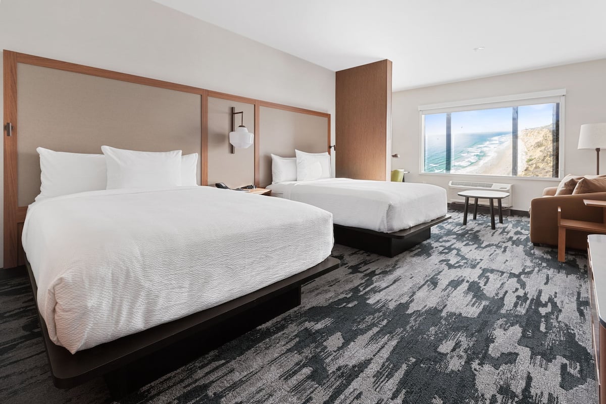 A hotel room with two beds, a small table, a chair, and a large window offering a view of the ocean.