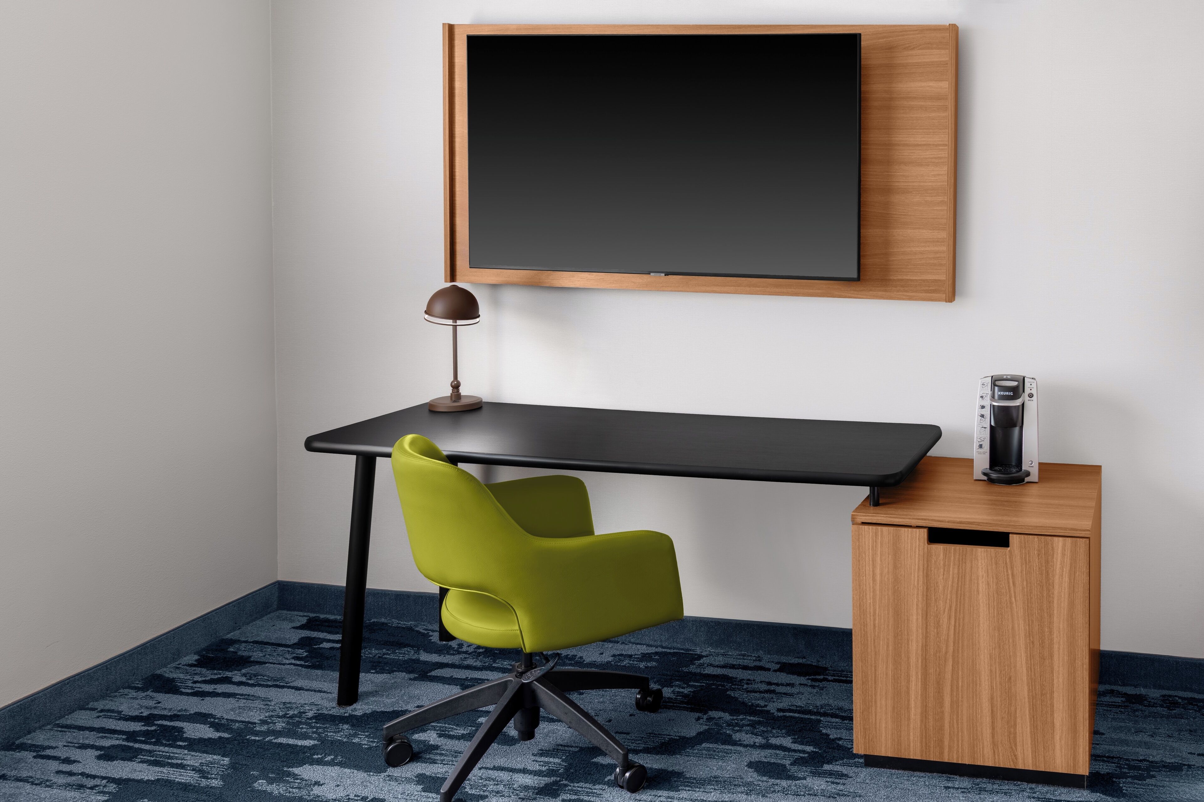 Un espace de bureau moderne avec un bureau noir, une chaise de bureau verte, une lampe noire et une machine à café posée sur une petite table d’appoint en bois.