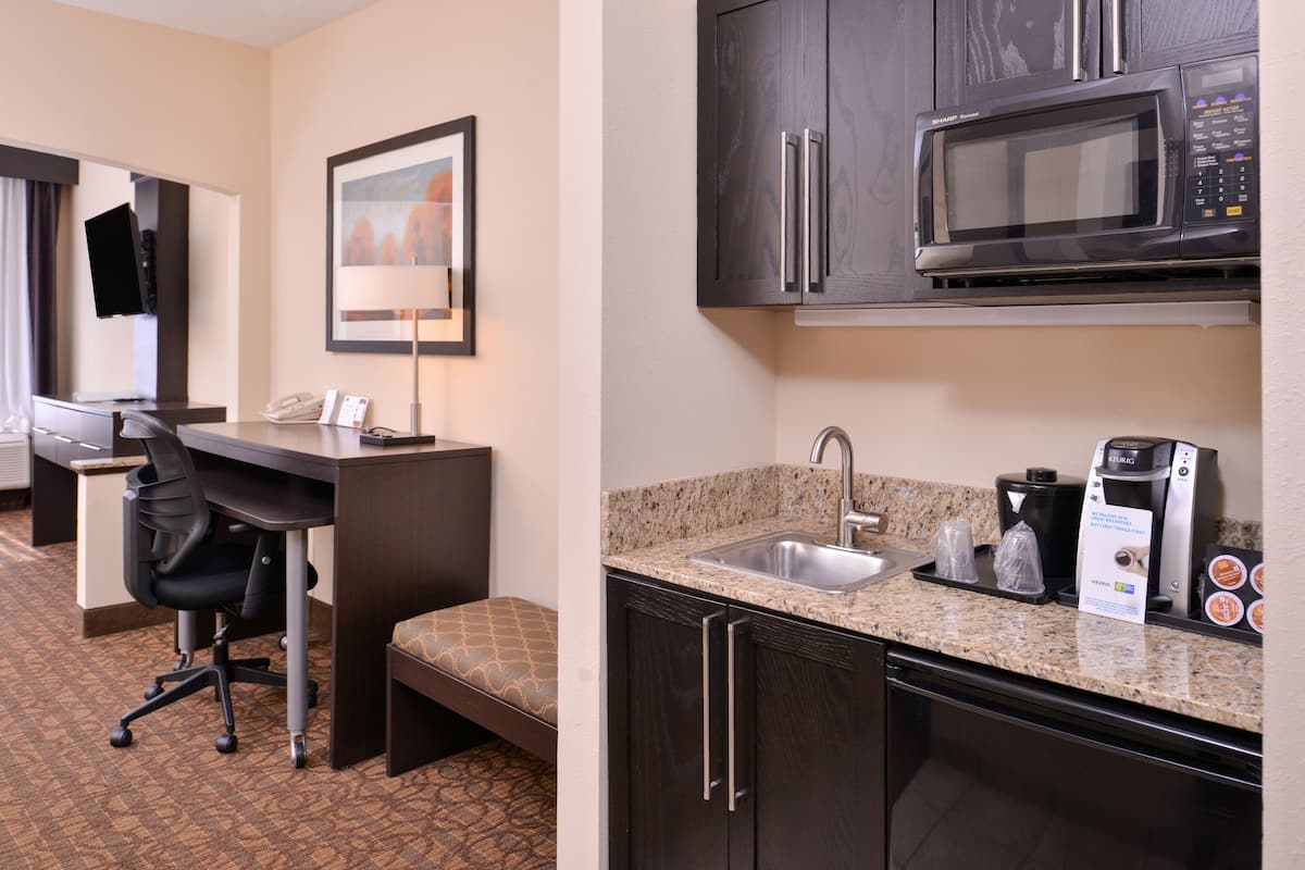 A hotel room with a desk, chair, and a kitchenette area including a microwave, sink, and coffee maker.