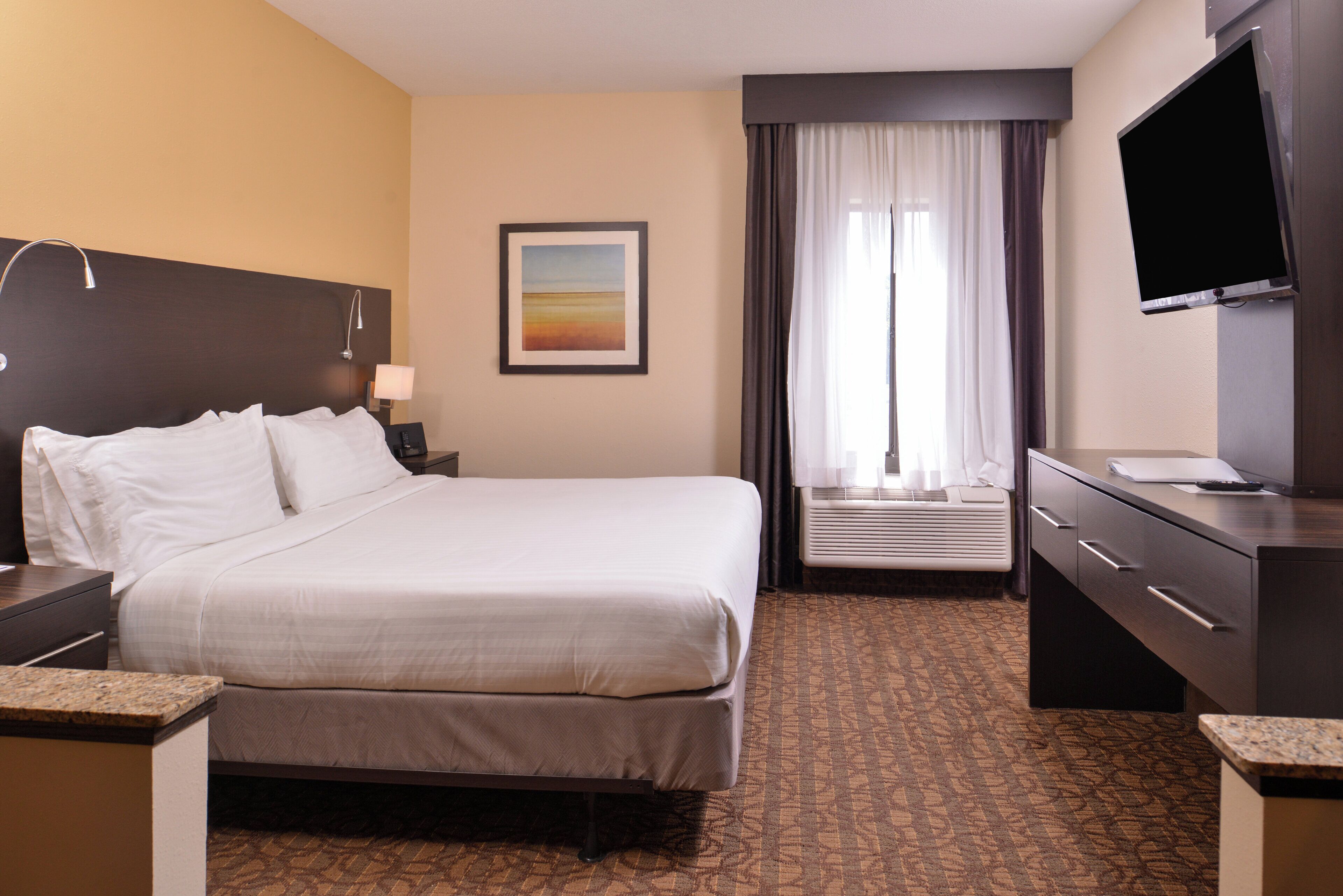 A hotel room with a bed, a desk, a television, a window with curtains, and a painting on the wall.
