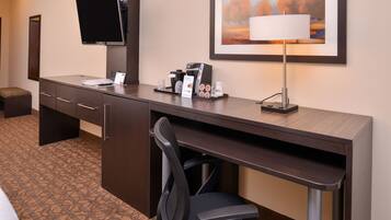 A hotel room with a desk, a chair, a flat-screen TV, a coffee maker, a lamp, and a framed picture on the wall.