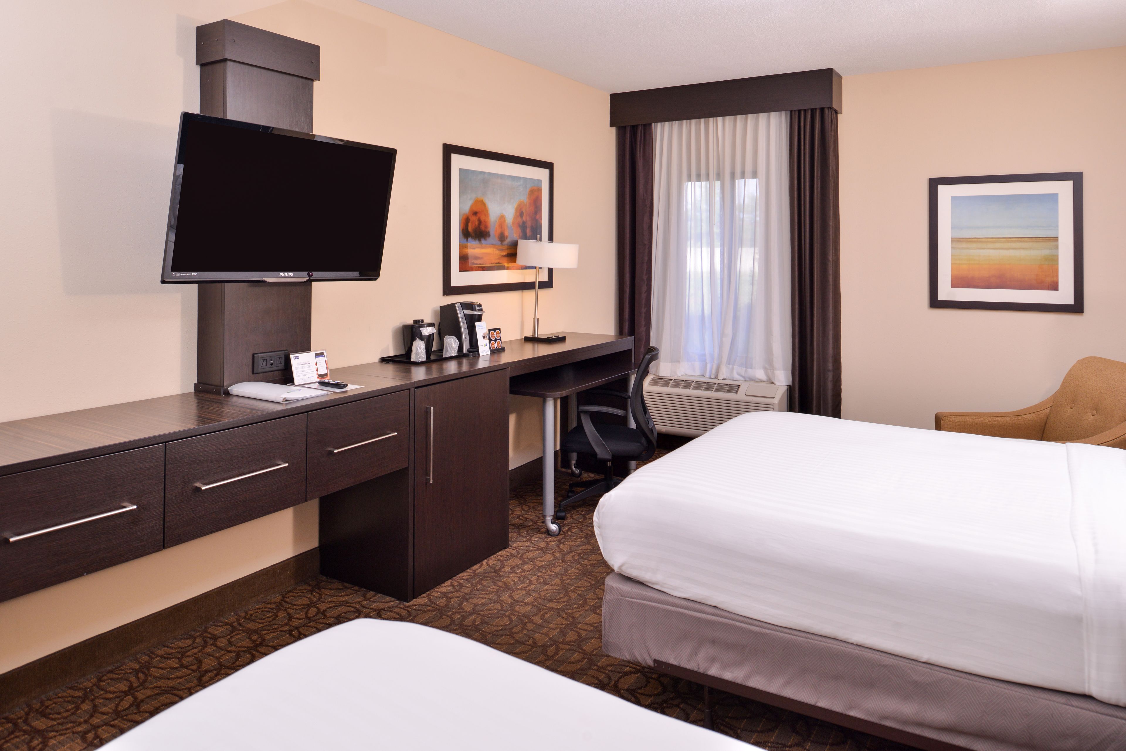 A hotel room with a bed, a desk with a flat-screen TV, a chair, and two framed pictures on the wall.