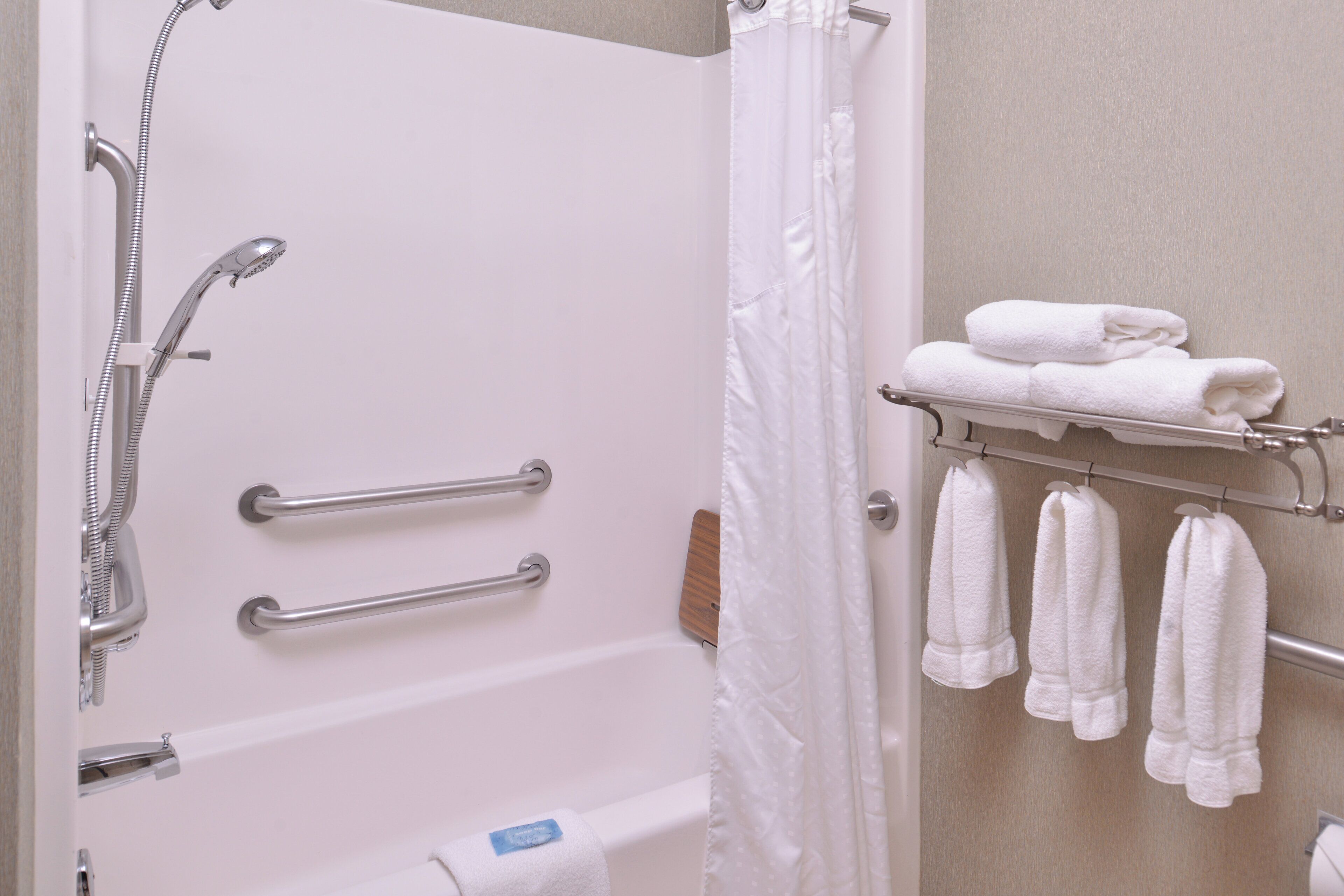 Un baño con ducha, barras de apoyo, un perchero con toallas y un dispensador de papel higiénico.