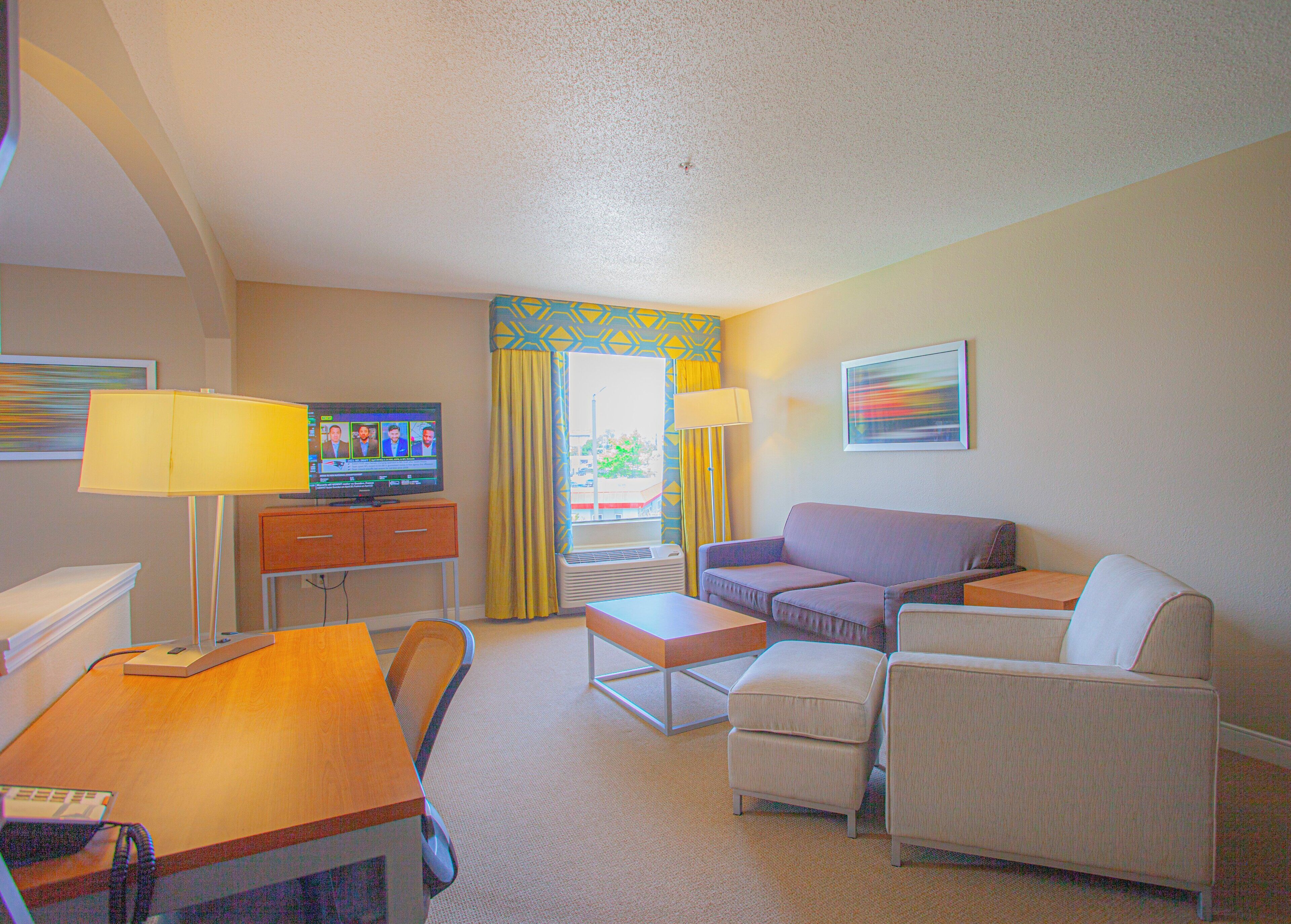 A hotel room with a desk, two chairs, a sofa, a television, and a window with curtains.