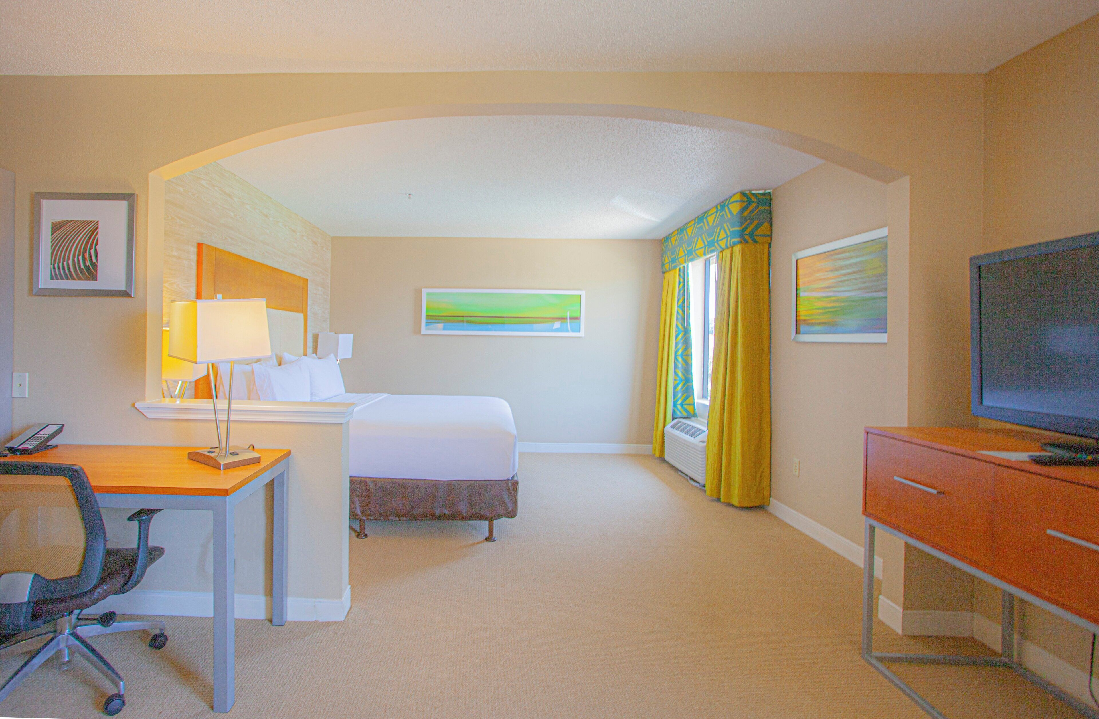 Une chambre d’hôtel équipée d’un lit, d’un bureau, d’une chaise, d’une télévision et décorée avec des œuvres d’art aux murs.