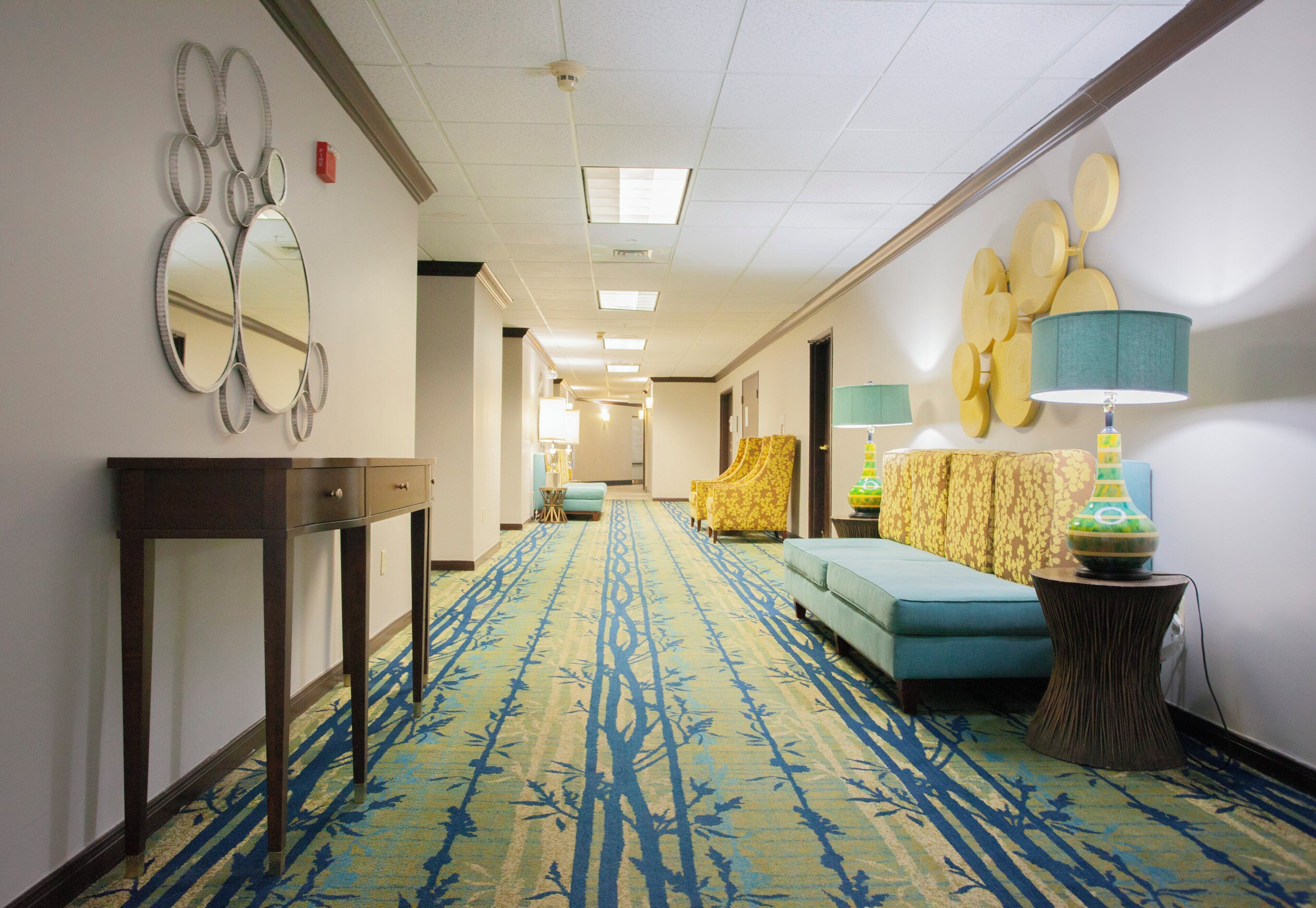 A hotel hallway with a patterned carpet, a sofa, a lamp, and a mirror on the wall.