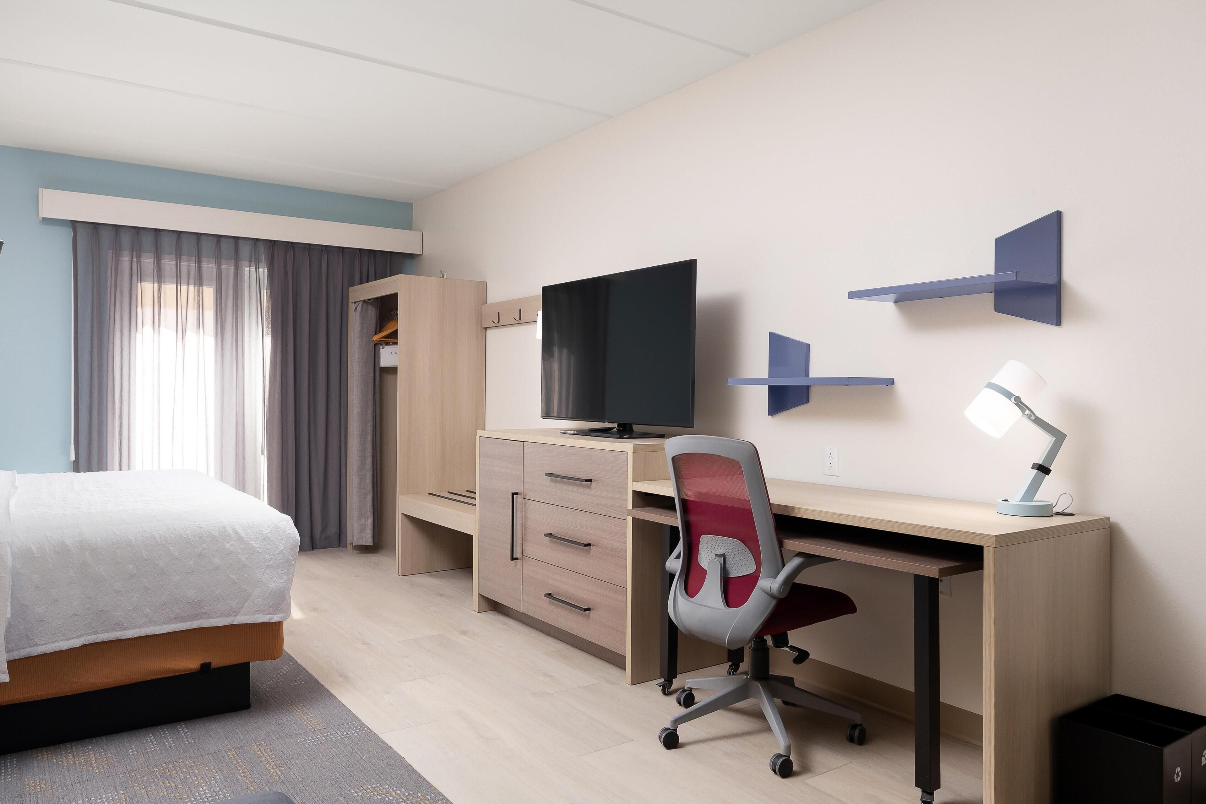 A modern hotel room with a bed, desk, chair, TV, and storage unit.