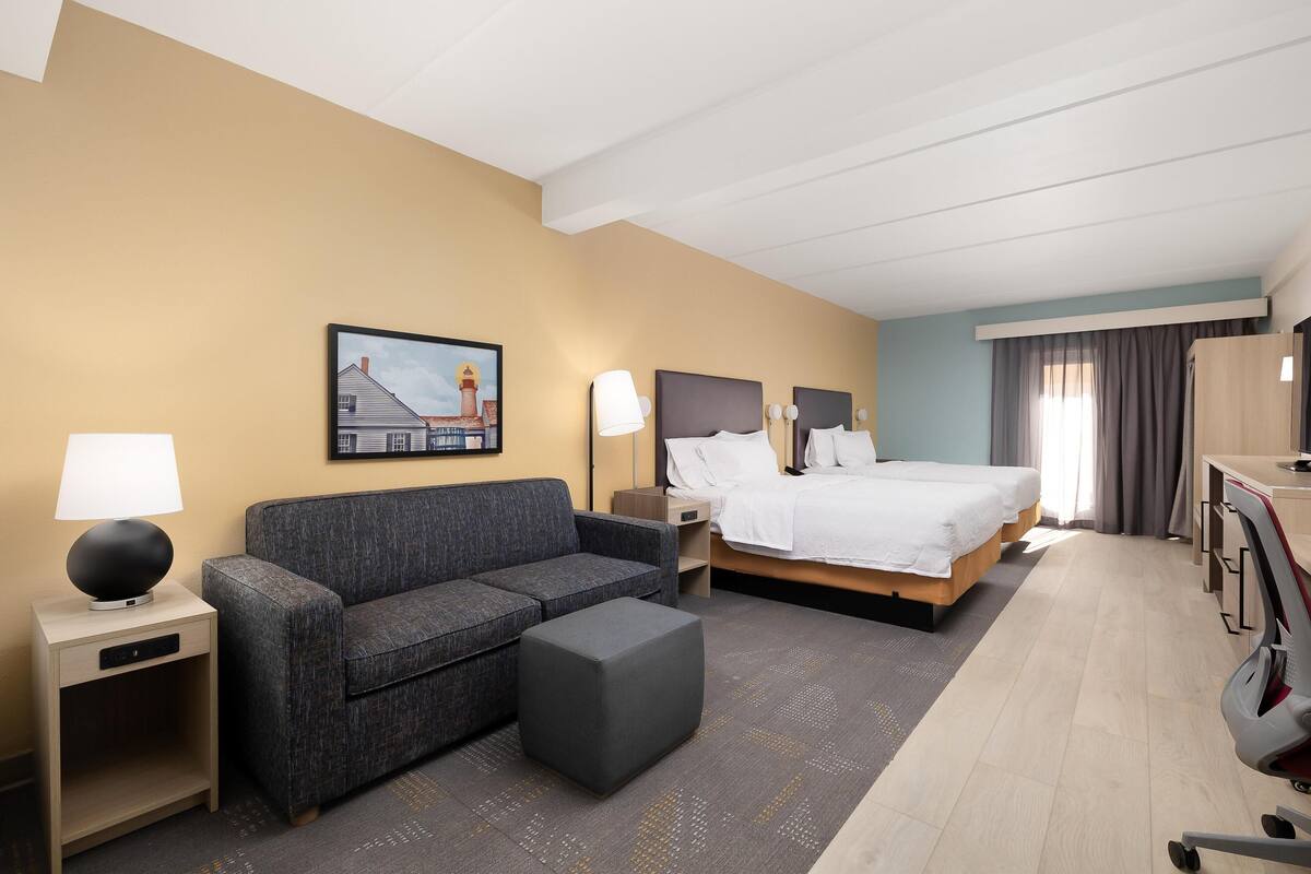 A hotel room with a bed, a sofa, a desk, a lamp, a nightstand, and a painting on the wall.