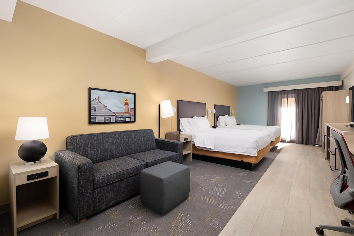 A hotel room with a bed, a sofa, a desk, a lamp, a nightstand, and a painting on the wall.