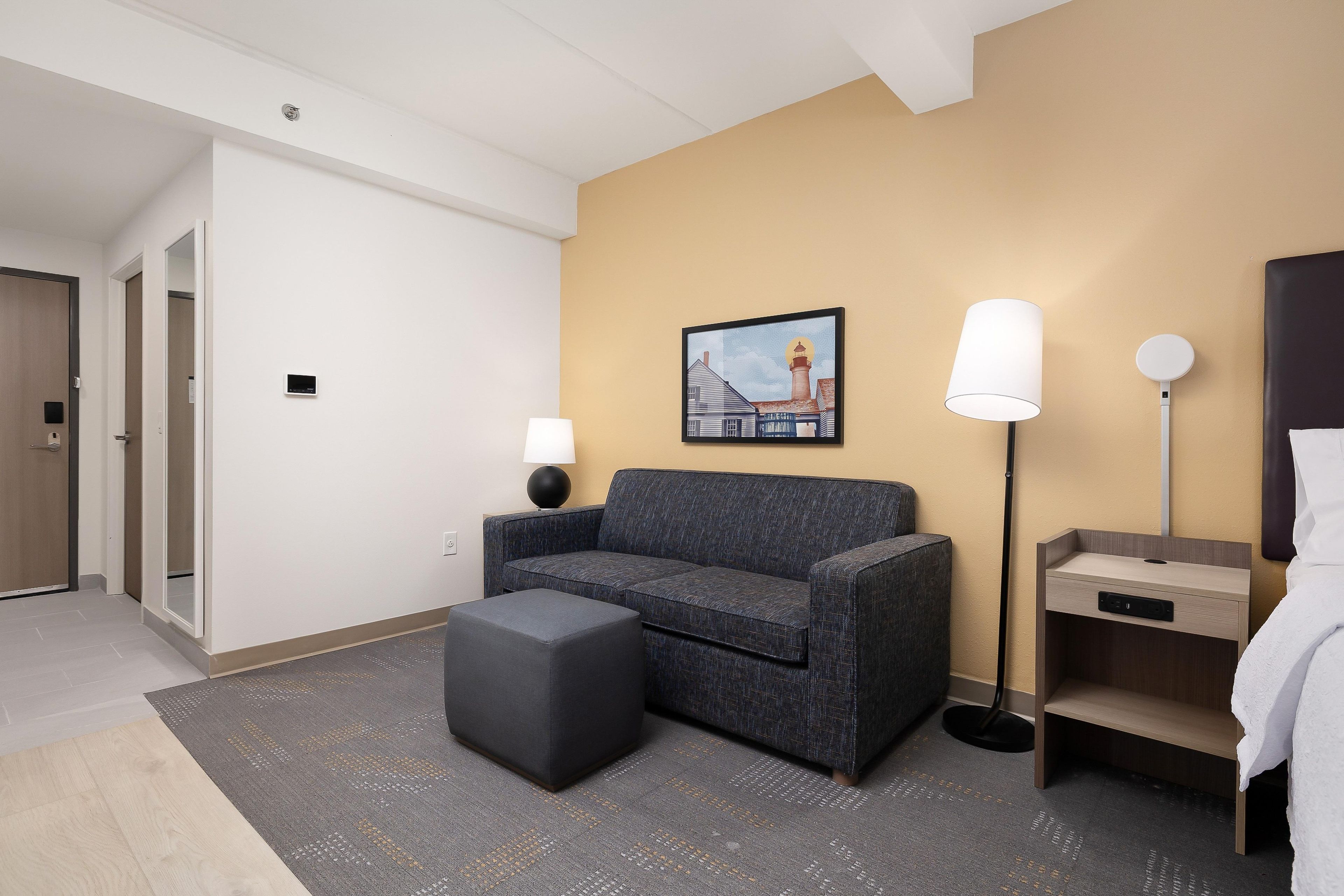 A hotel room with a sofa, a small table, a lamp, a painting, and a bedside table with a lamp.