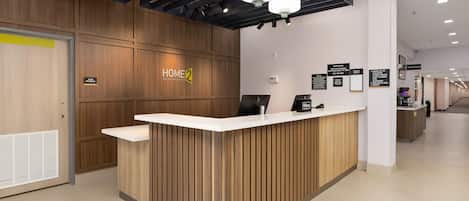 A hotel reception desk with a wooden finish, a digital screen, and a sign indicating 'Home 2 Suites by Hilton.'