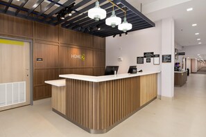 Ein Hotelrezeptionstresen mit Holzoptik, einem digitalen Bildschirm und einem Schild mit der Aufschrift 'Home 2 Suites by Hilton'.
