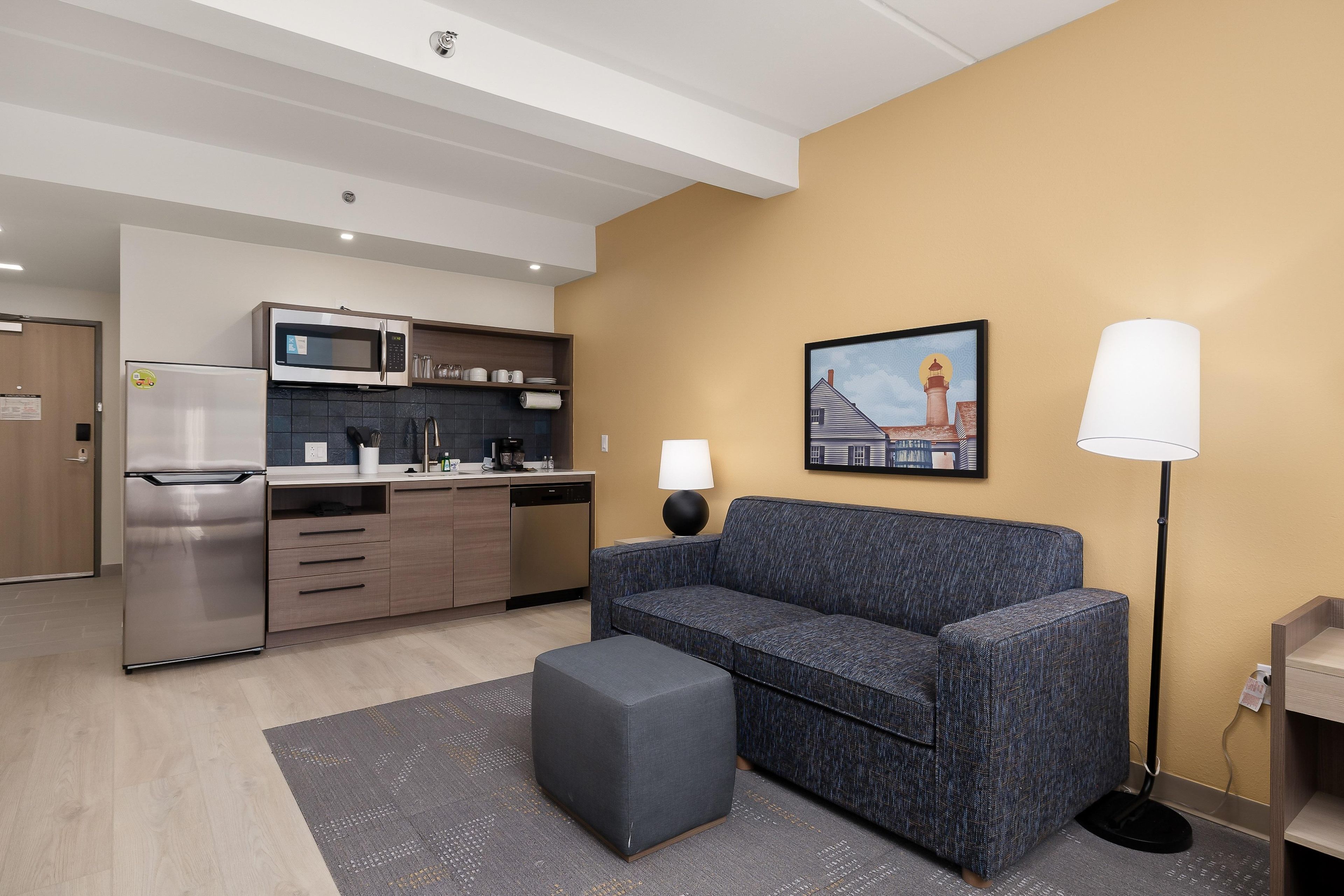 A modern kitchenette with a microwave, refrigerator, and dishwasher. There is a gray sofa with a matching ottoman and a floor lamp. A painting of a lighthouse is on the wall.
