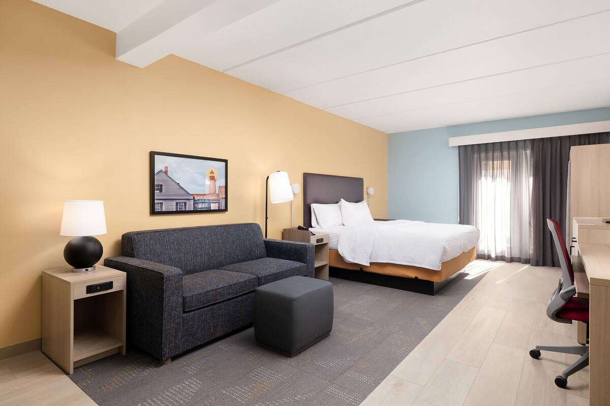 A hotel room with a bed, a sofa, a desk, a lamp, and a painting on the wall.