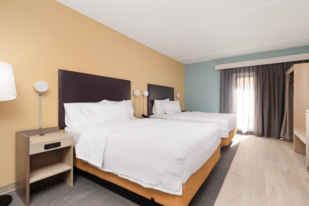 A hotel room with two beds, a bedside table with a lamp, a wardrobe, and a window with curtains.