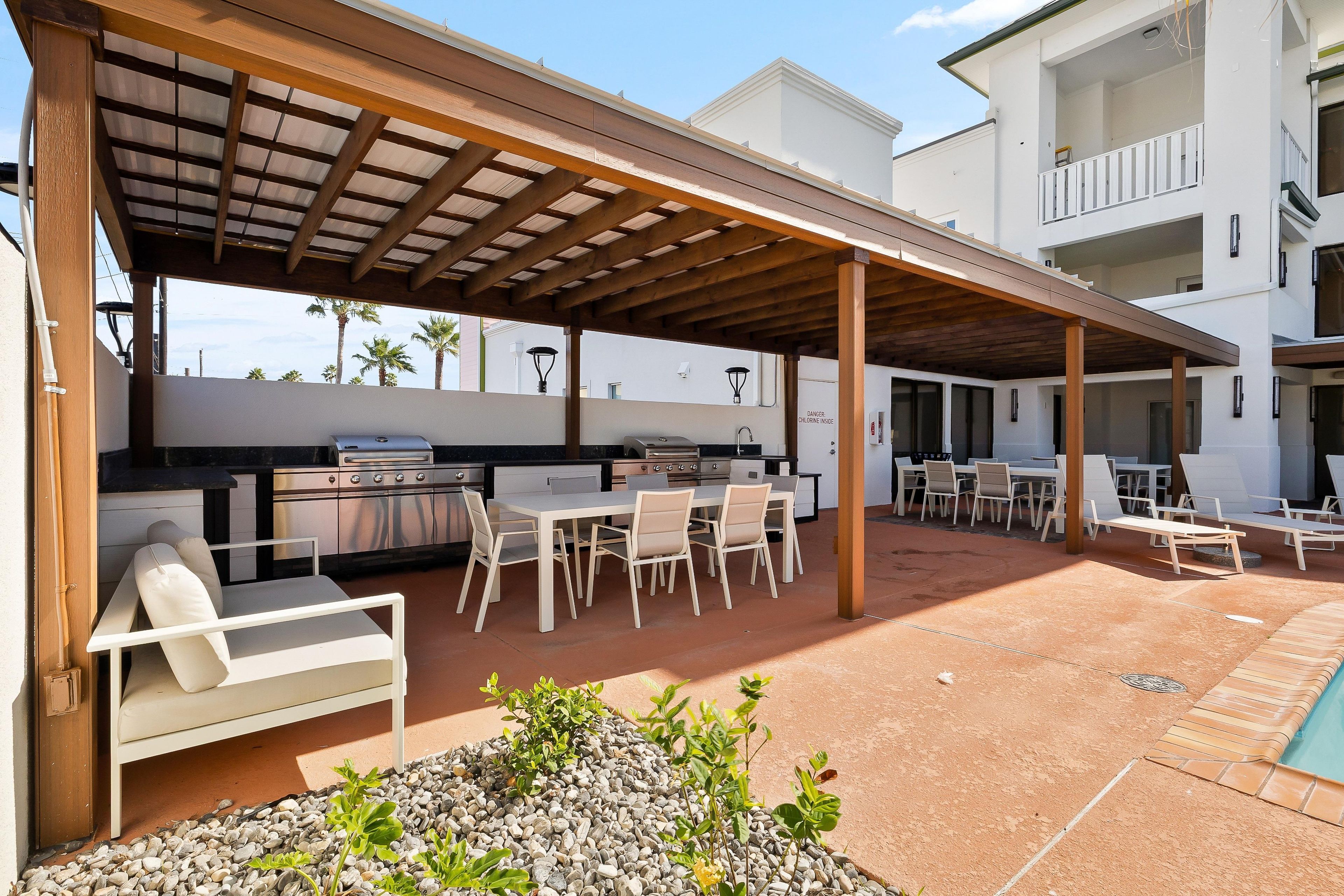 Un patio exterior con zona de parrilla cubierta, asientos y una piscina.