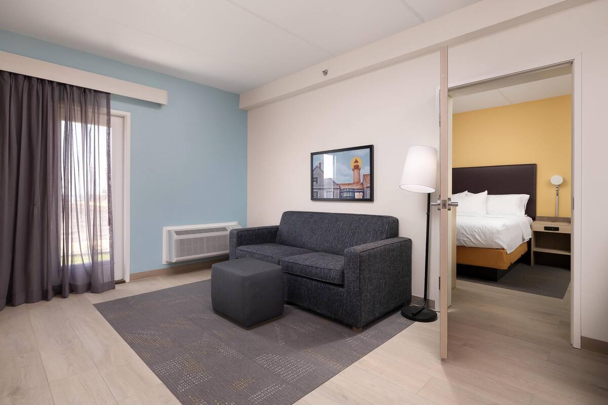 A hotel room with a sofa, ottoman, a framed picture on the wall, a lamp, and a bed with a headboard.