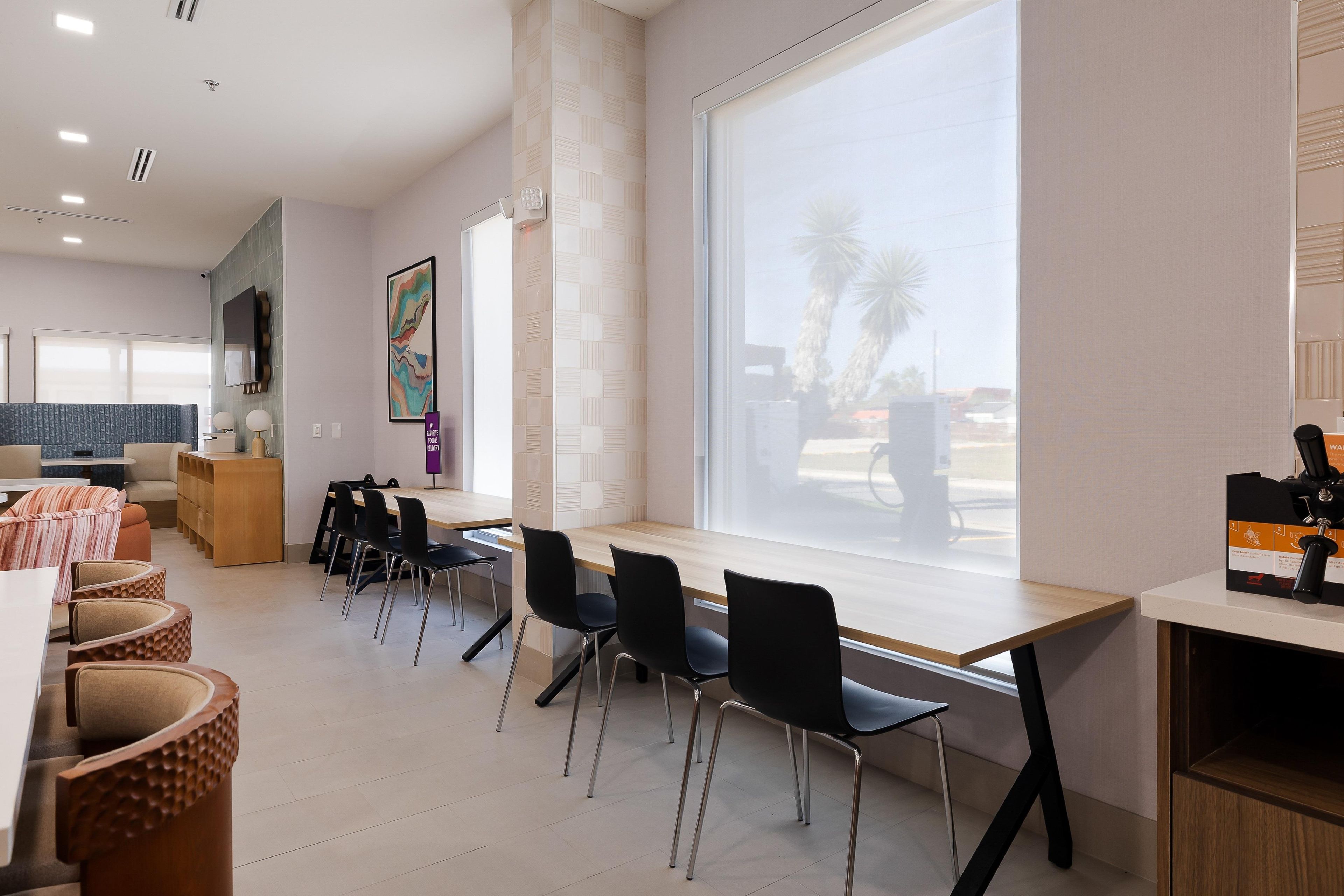 A modern dining area with a long table, black chairs, and a large window with a view of palm trees.