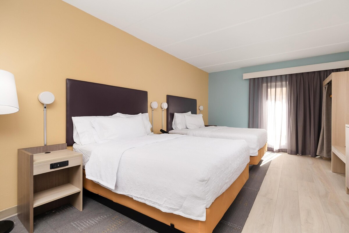 A hotel room with two beds, a bedside table with a lamp, a wardrobe, and a window with curtains.