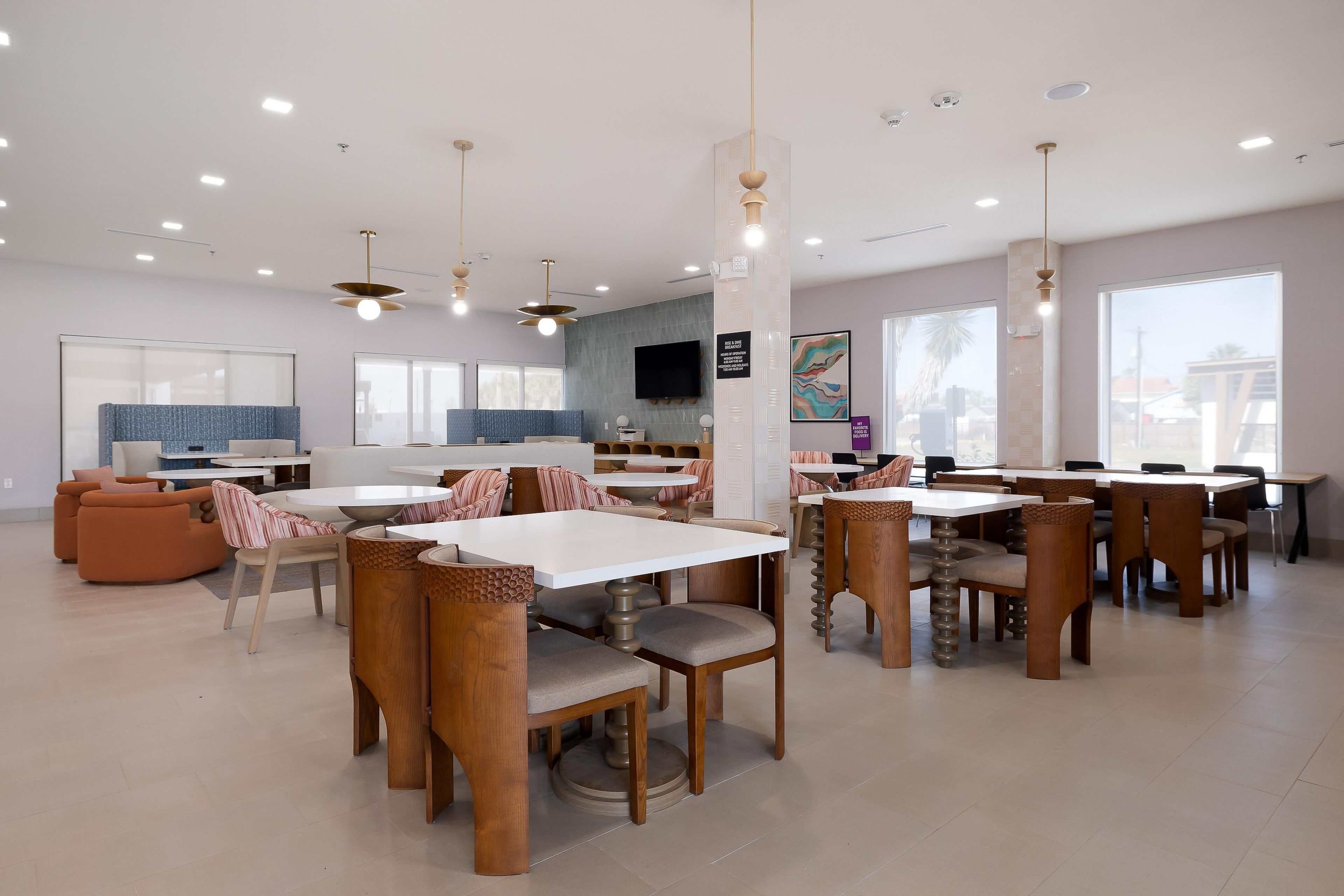 A modern dining area with multiple tables and chairs, a kitchen in the background, and large windows.