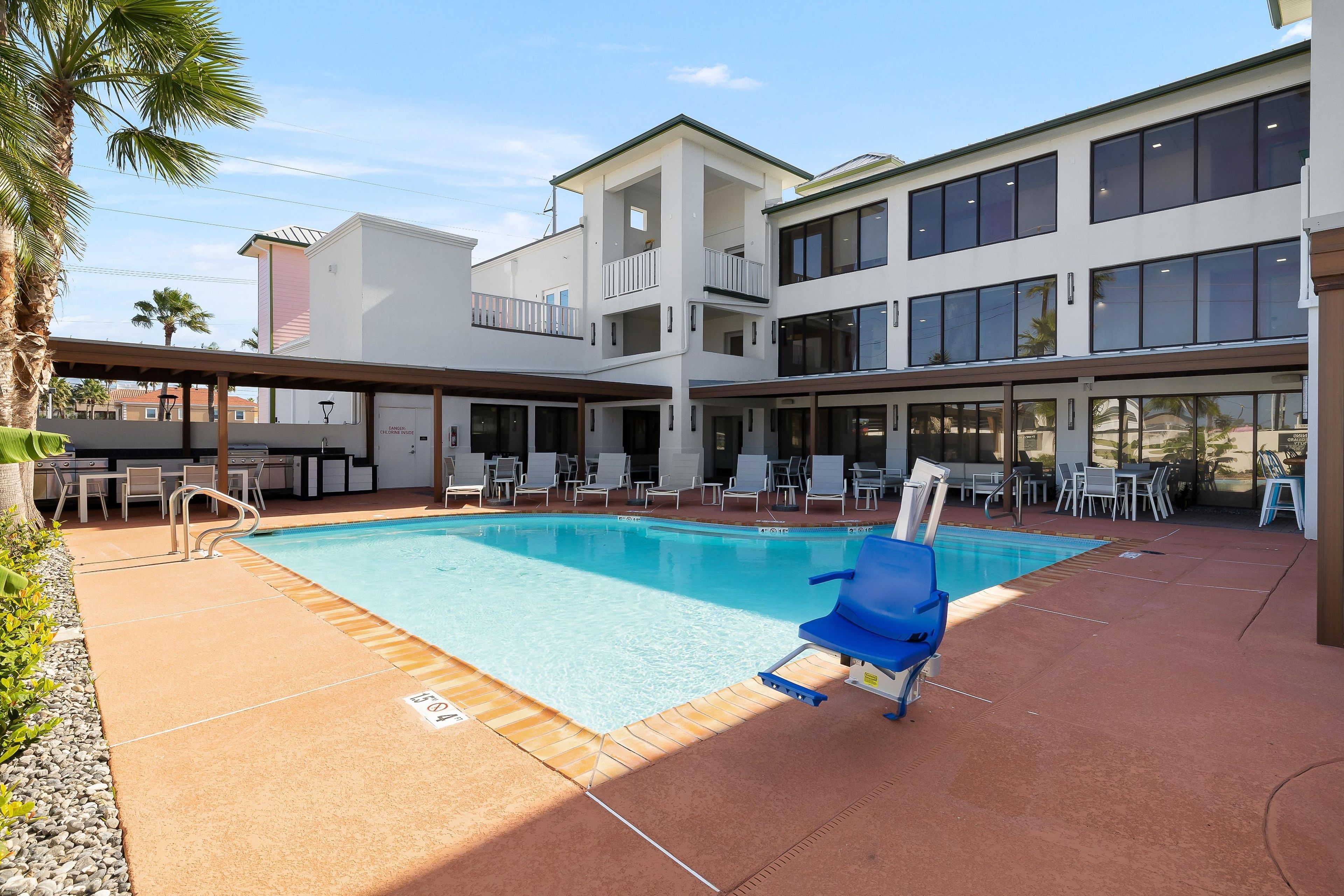A pool area with lounge chairs, a barbecue setup, and a two-story building with large windows.