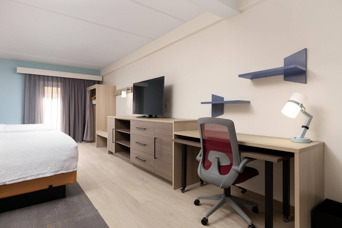 A hotel room with a bed, a desk with a chair, a TV, and a lamp.
