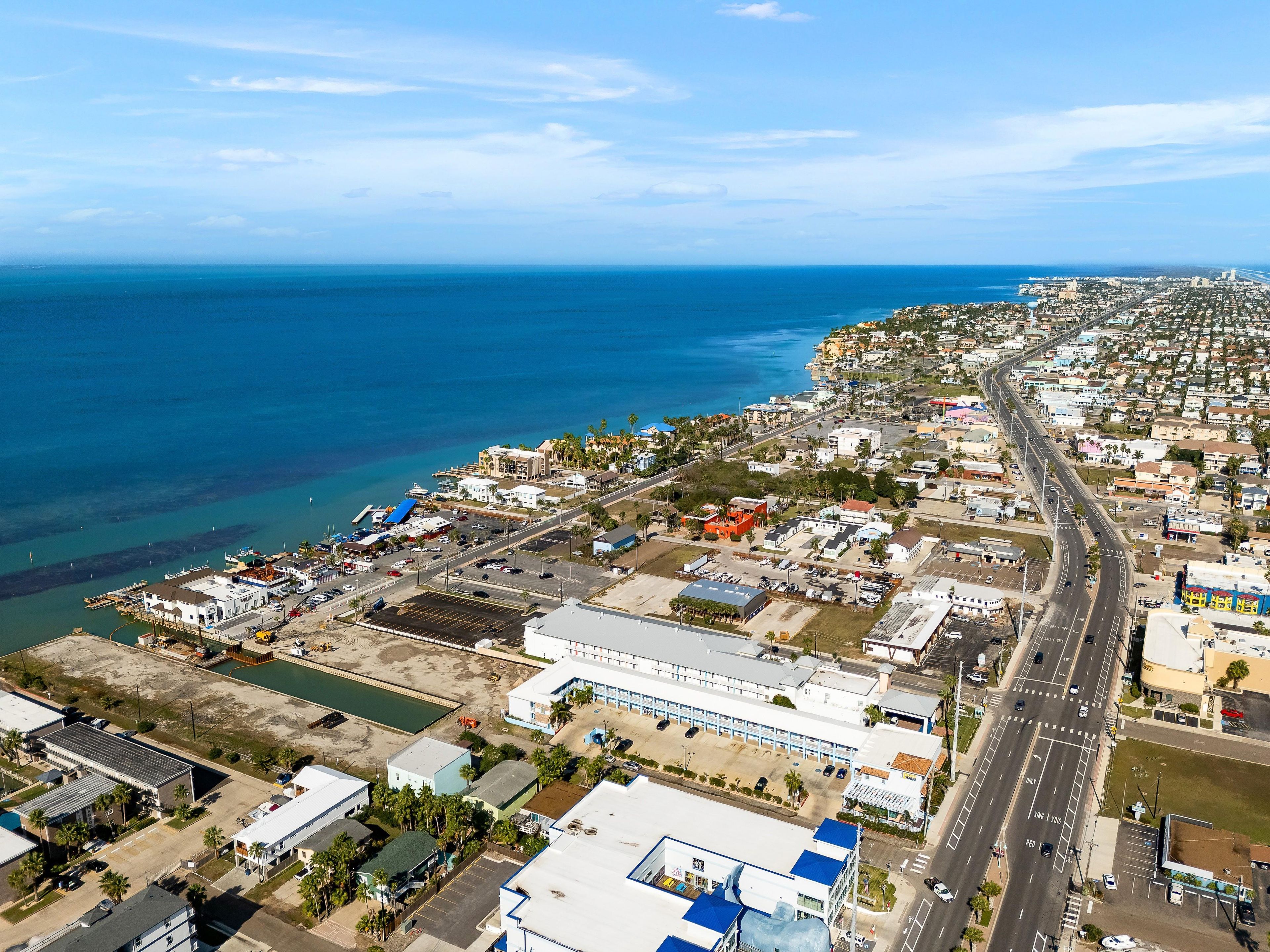 An aerial view of a coastal city with a prominent road running through it, a marina, and various buildings.