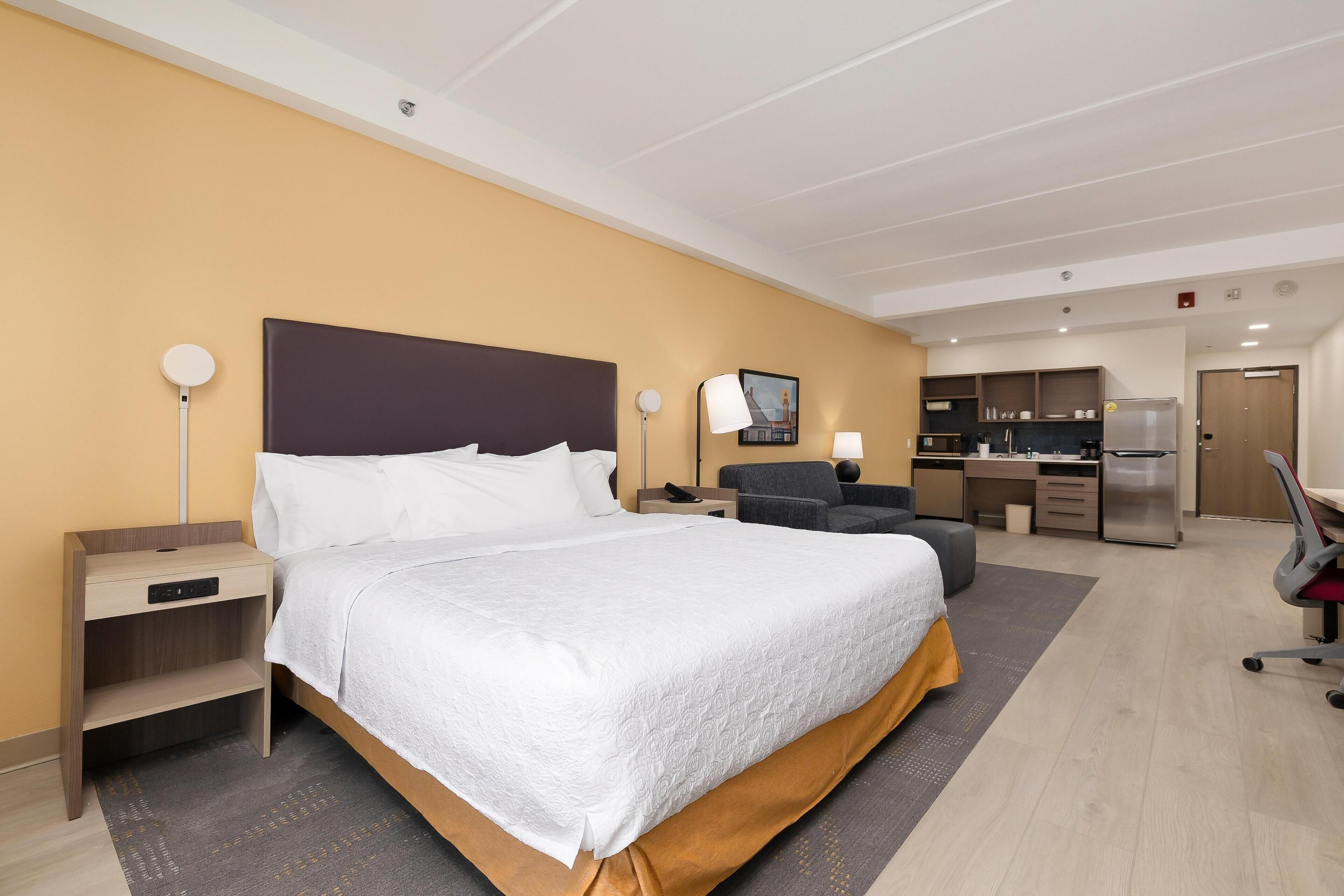 A hotel room with a bed, bedside tables, a desk, a chair, a lamp, a refrigerator, and a kitchen area.