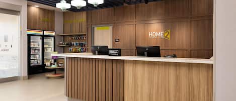 Un mostrador de recepción de hotel con detalles de madera, un cartel que dice 'HOME 2' y una nevera con varios artículos.