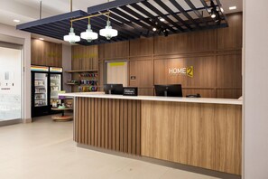 A hotel reception desk with a wooden finish, a sign reading 'HOME 2', and a minibar with various items.