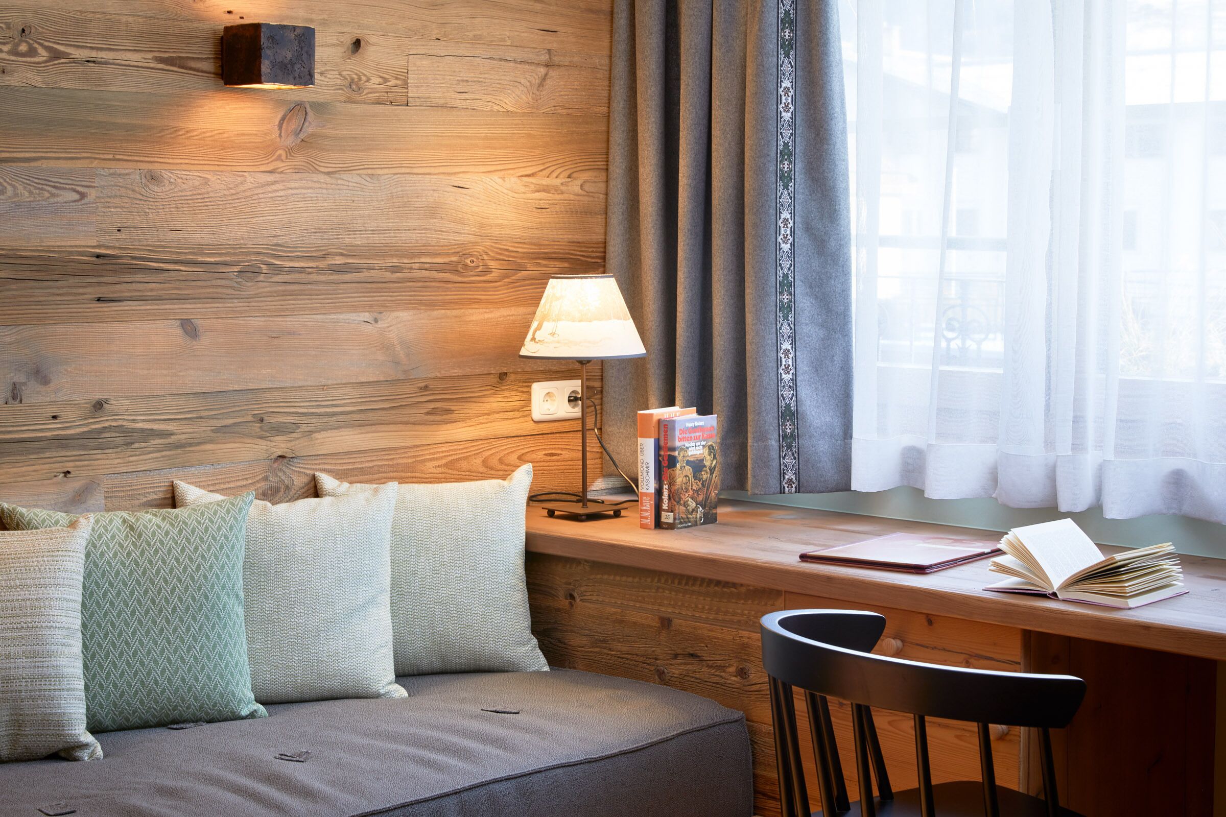 A cozy room with a wooden wall, a desk with books, a lamp, and a window with curtains.
