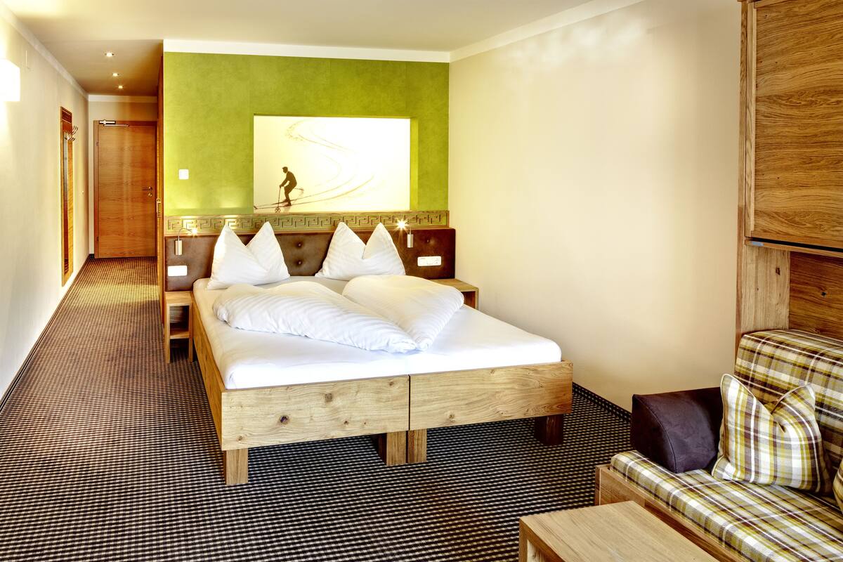 Une chambre d’hôtel avec un grand lit, une tête de lit en bois, un mur d’accent vert, un canapé et un sol à motifs.