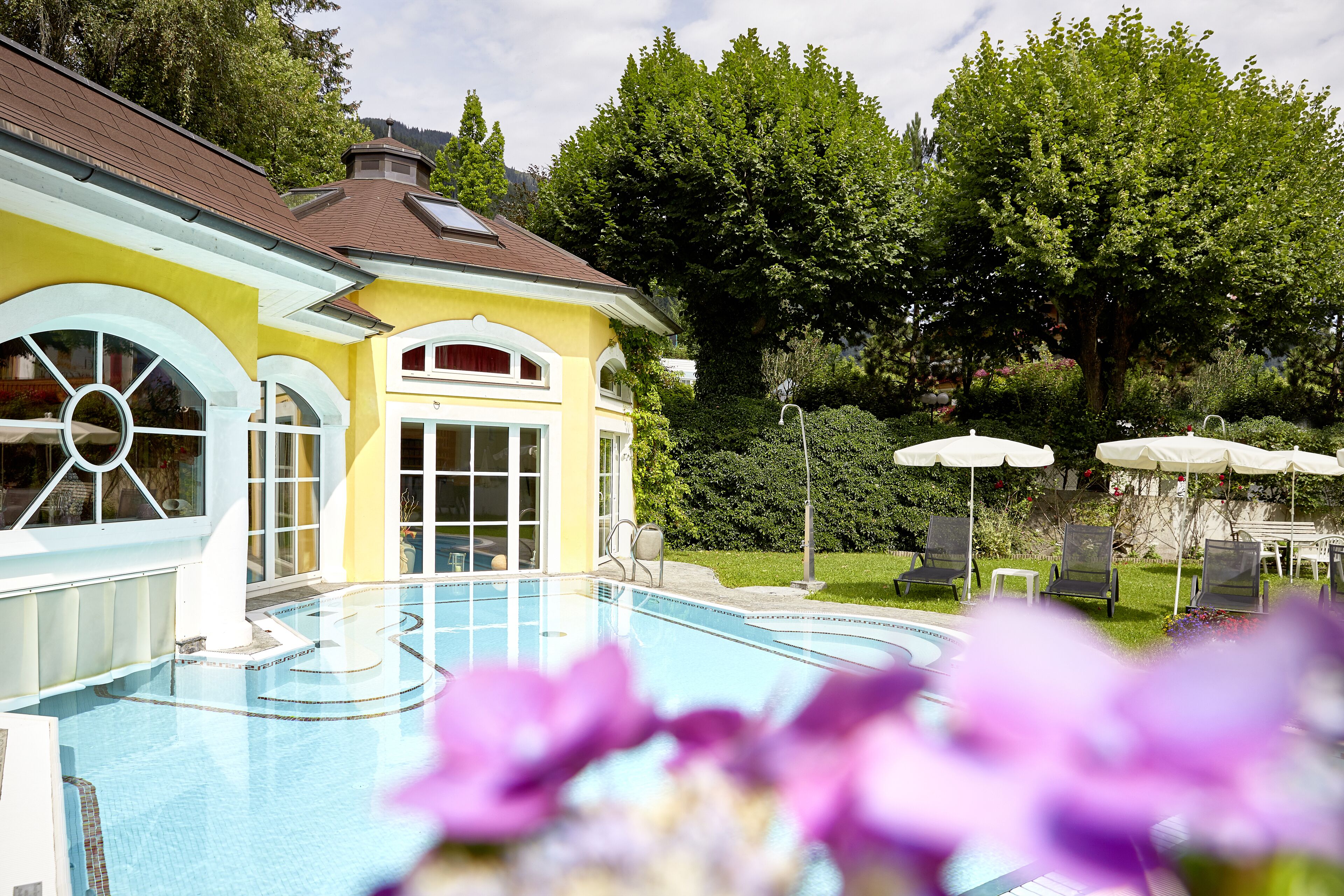 A swimming pool with a yellow building in the background, surrounded by greenery and outdoor seating.