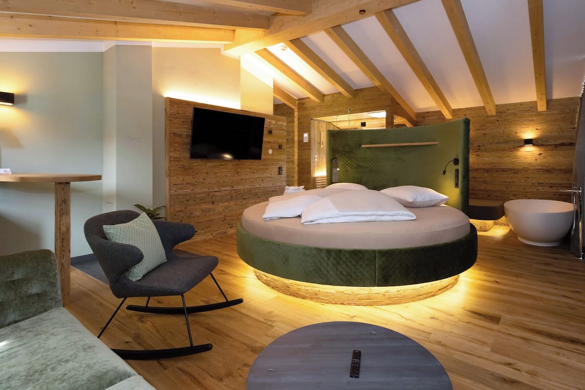 Un lit rond avec une tête de lit verte, un téléviseur à écran plat fixé à un mur en bois, un fauteuil à bascule et une petite table avec une télécommande.