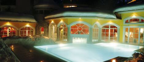 A hotel with a large outdoor pool surrounded by snow, featuring a stone facade and arched windows.