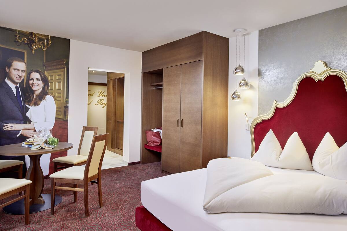 Une chambre d’hôtel avec un grand lit, une armoire en bois, une table à manger avec des chaises et un mur orné d’un tableau encadré.