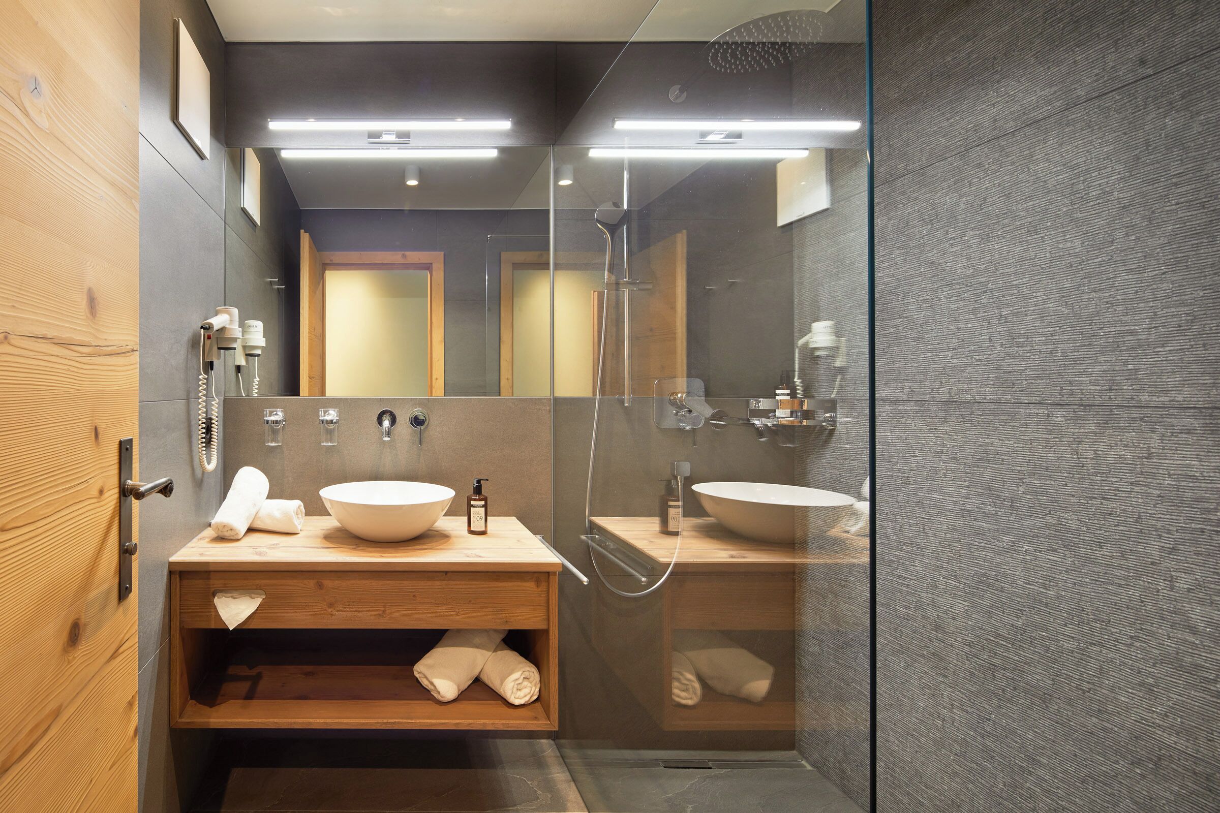 A modern bathroom with a wooden vanity, two sinks, a glass shower enclosure, and a large mirror.