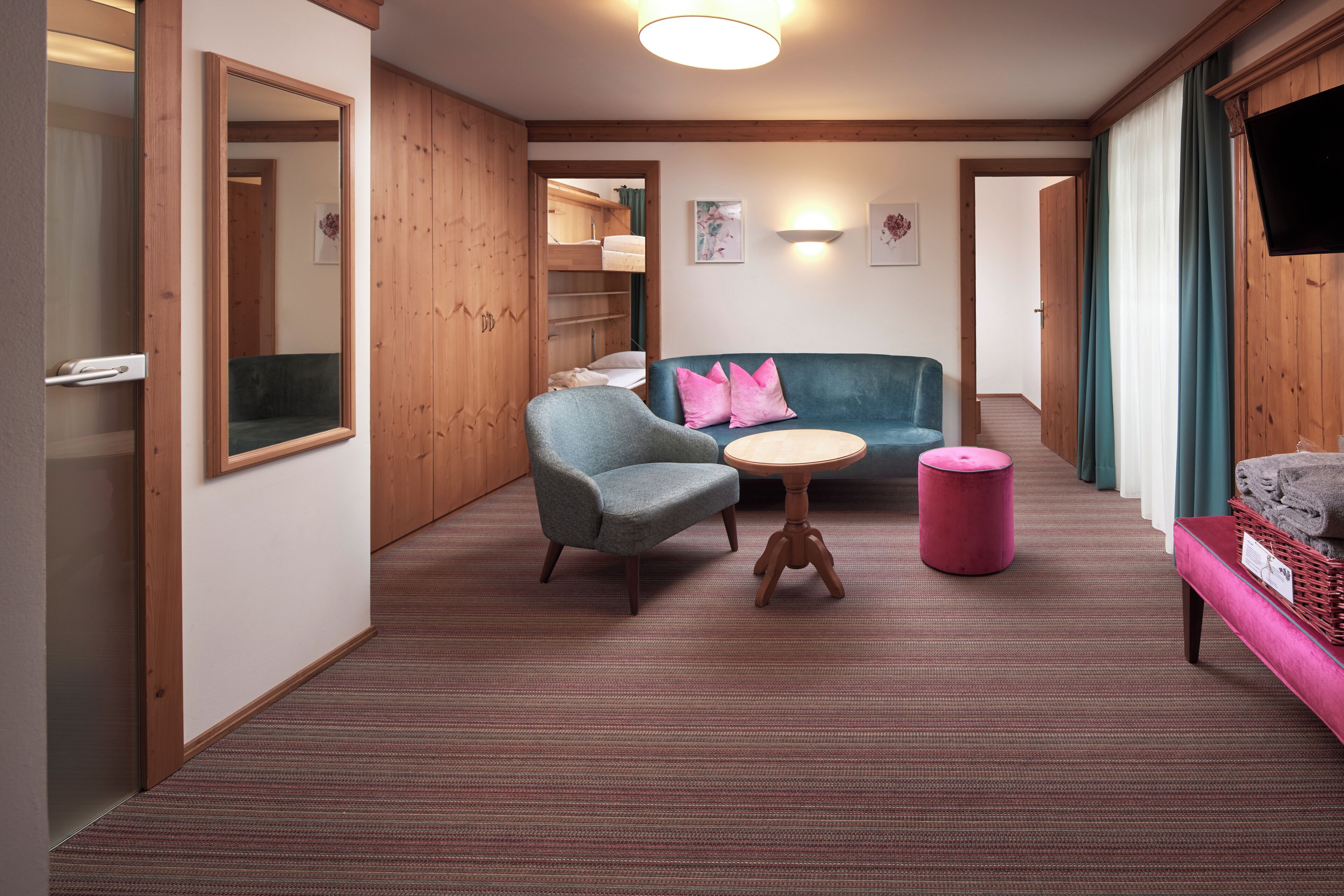 Een hotelkamer met een houten vloer, een bank, een stoel, een klein rond tafeltje, een roze poef en een televisie aan de muur.