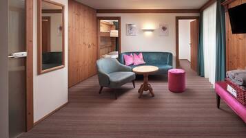 Een hotelkamer met een houten vloer, een bank, een stoel, een klein rond tafeltje, een roze poef en een televisie aan de muur.