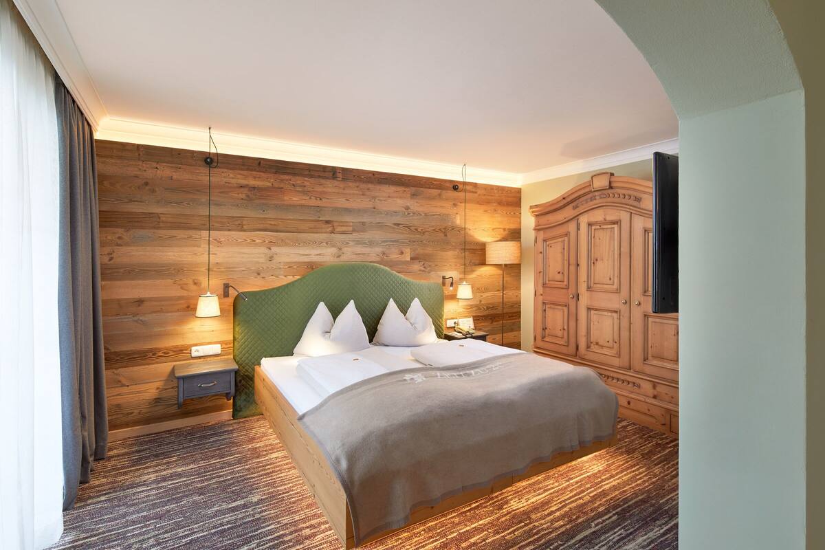 Une chambre d’hôtel avec un mur en bois, une tête de lit recouverte d’un tissu vert, un grand lit avec du linge de lit blanc, deux lampes de chevet, une table de chevet et une armoire en bois.